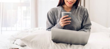 Genç gülümseyen mutlu Asyalı kadın dizüstü bilgisayar kullanarak rahatlıyor ve yatak odasında kahve içiyor. Kız, tatil sabahında evde yatağında defter ile ders çalışıyor. 