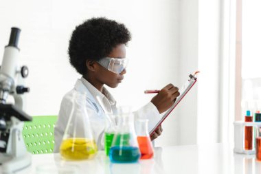 Afrika kökenli, sevimli, küçük bir çocuk araştırma öğreniyor ve kimyasal bir deney yaparken fen bilimleri sınıfında sıvıyı analiz edip karıştırıyor. Eğitim ve bilim konsepti.