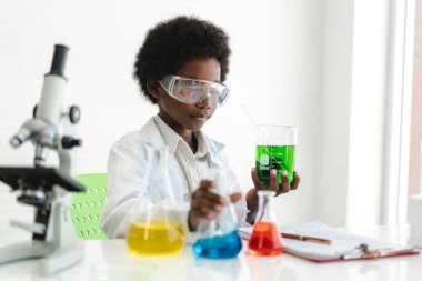 Afrika kökenli, sevimli, küçük bir çocuk araştırma öğreniyor ve kimyasal bir deney yaparken fen bilimleri sınıfında sıvıyı analiz edip karıştırıyor. Eğitim ve bilim konsepti.