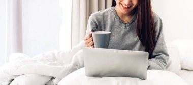 Genç gülümseyen mutlu Asyalı kadın dizüstü bilgisayar kullanarak rahatlıyor ve evdeki yatak odasında kahve içiyor. Genç yaratıcı kız klavyede çalışıyor ve daktilo kullanıyor. Ev konseptinden çalışıyor.