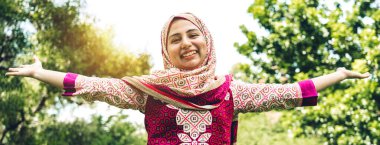 Mutlu Asyalı güzel Müslüman kadın portresi tesettürlü ayakta dinleniyor kollarını açıyor ve yeşil park arka planında temiz havanın tadını çıkarıyor.