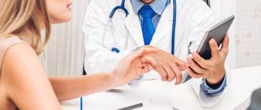 Maskeli, üniformalı, steteskop hizmeti veren bir erkek doktor hastane kontrolleri hakkında maske takan hasta kadın hastalarla konuşma ve danışmanlık konusunda yardımcı oluyor.