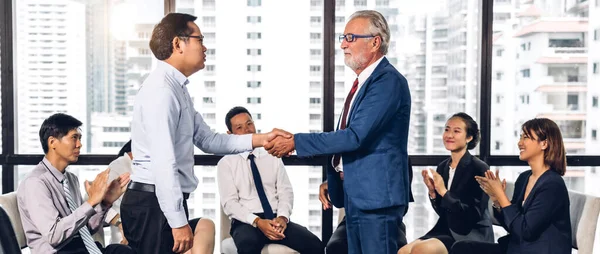 在现代办公室里 两个穿着雅致西装的商业伙伴在一群随意拍手的同事面前成功地握手 — 图库照片