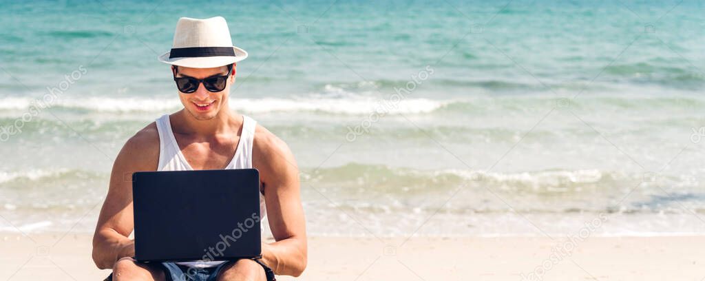 Portrait of smiling happy handsome sexy man relax in sunglasses and straw hat working on his laptop computer on the tropical beach.Summer vacations and travel