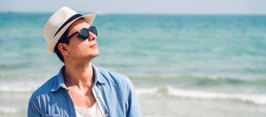 Gülümseyen yakışıklı erkek modelin portresi, şık güneş gözlükleriyle ve hipster yaz hasır şapkasıyla tropik sahilde durup Camara 'ya bakarken keyif çatıyor ve rahatlıyor.
