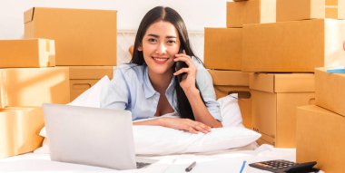 Genç, gülümseyen, güzel sahibi Asyalı kadın serbest meslek edinen, online alışveriş yapan, dizüstü bilgisayarda evde bir paket kutusuyla çalışan KOBİ işi online ve teslimat konsepti.