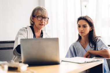 Steteskoplu üniforma giyen kıdemli kadın doktor hasta kadın hastalarla hastanede dizüstü bilgisayarla yapılan kontroller hakkında konuşmak ve danışmanlık yapmak için yardımcı oluyor.