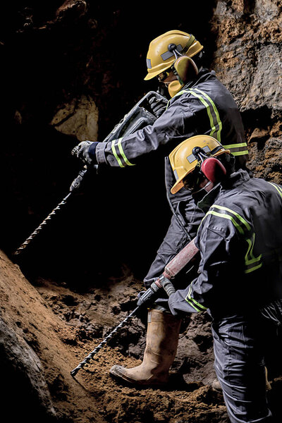 Two workers with helmet and protective suit using drill-machines in an underground environment