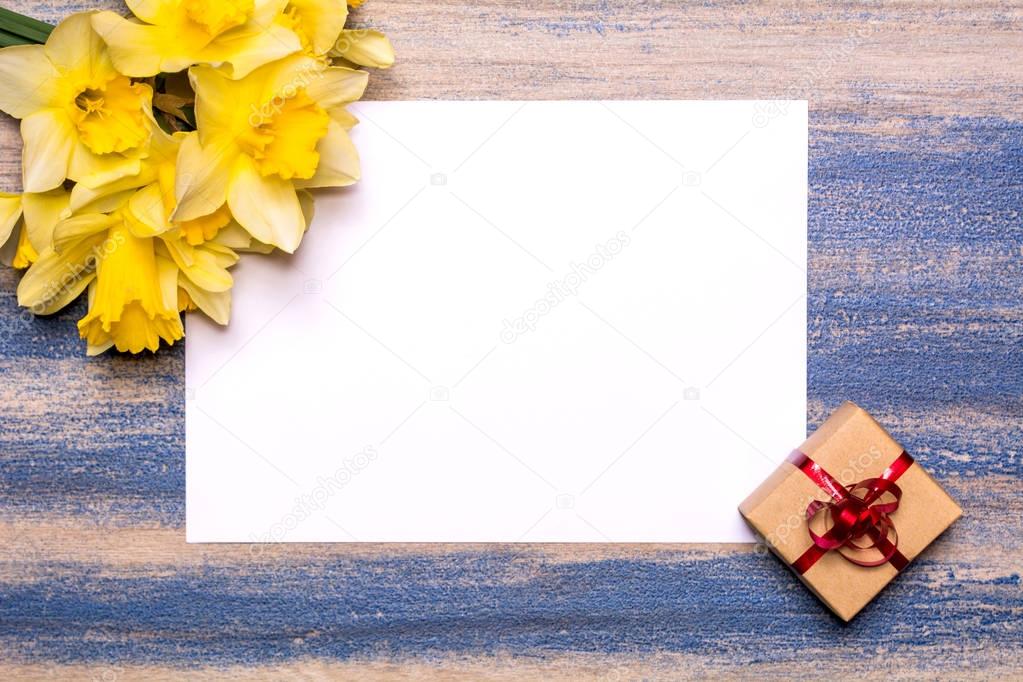 A bouquet of daffodils, a gift with a red ribbon and a sheet of white paper on a wooden background.