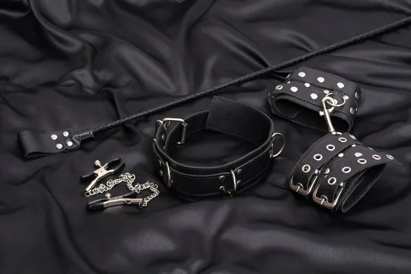 Leather handcuffs, nipple clamps, chain collar and stack on black silk background. Accessories for adult sexual game. BDSM outfit. — Stock Photo, Image