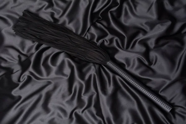 Látigo negro sobre un fondo de seda negro. Accesorios para juegos sexuales para adultos. Juguetes para BDSM —  Fotos de Stock