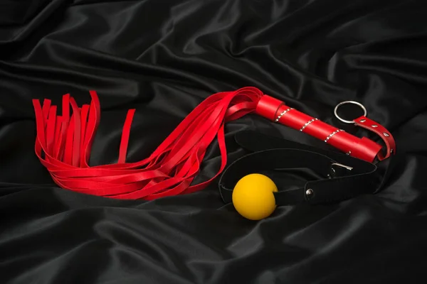 Red whip and gag on a black silk background. Accessories for adult sexual games.