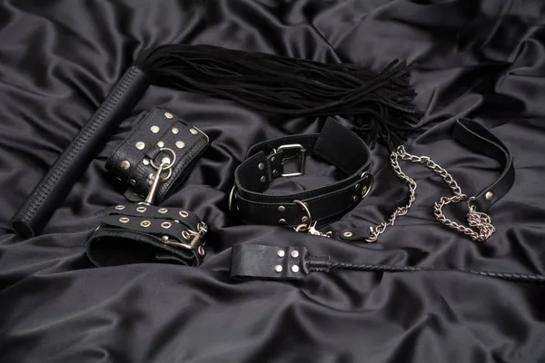 Leather handcuffs, black whip, chain collar and stack on black background. Accessories for adult sexual game. BDSM outfit.