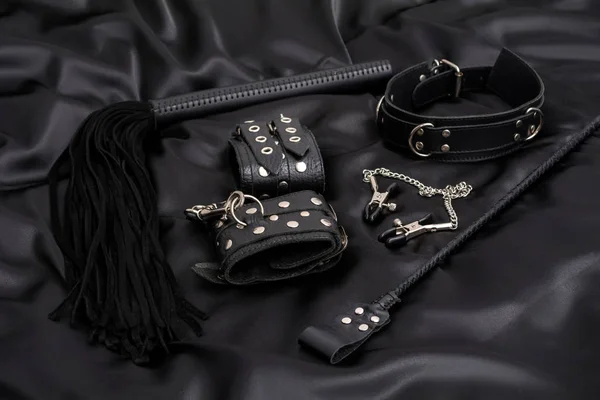 Leather handcuffs, black whip, chain collar, nipple clamps and stack on black background. BDSM outfit.