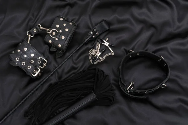 Leather handcuffs, black whip, chain collar, nipple clamps and stack on black background. BDSM outfit.
