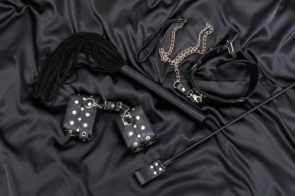 Leather handcuffs, black whip, chain collar and stack on black background. Accessories for adult sexual game. BDSM outfit.