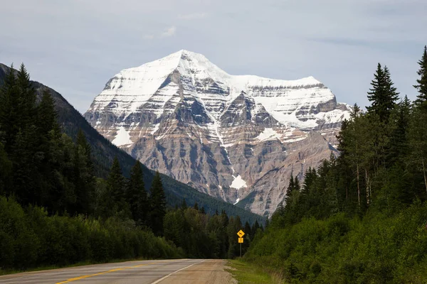 Mountain road passing through evergreen forests below the towering snow covered peak of below Mount Robson, British Columbia, Canada on a hazy day in a picturesque landscape