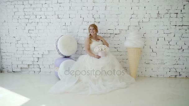 Niedliche junge blonde Frau im Hochzeitskleid mit Blumenstrauß posiert im Studio auf dem Hintergrund riesiges Eis und Makkaroni — Stockvideo
