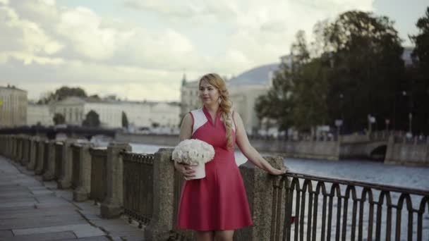 Niedliche junge blonde Frau in rotem Kleid, die mit Blumen auf der Straße spaziert — Stockvideo