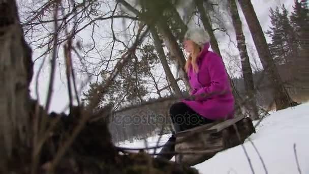 Giovane donna bionda in cappotto rosa seduta a panchina nella foresta invernale — Video Stock