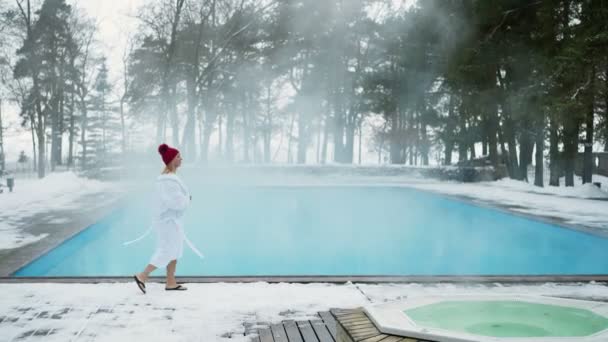 Mujer rubia joven en albornoz y cabaña roja cerca de la piscina al aire libre en invierno — Vídeos de Stock