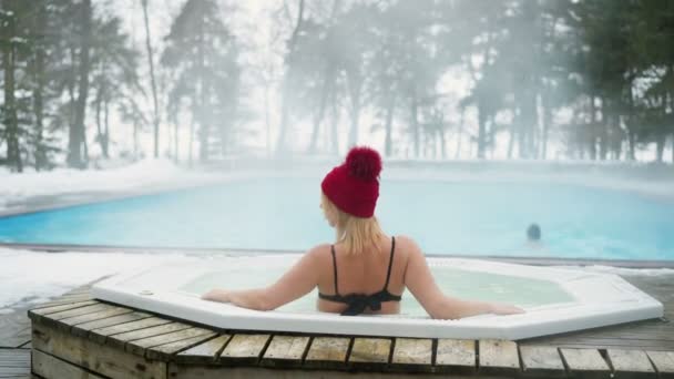 Nuori blondi nainen punaisessa mökissä kylpyammeessa porealtaassa ulkona talvella — kuvapankkivideo