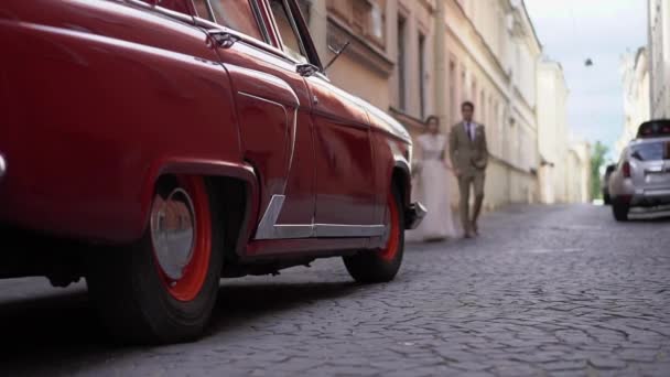 Νεαρό ζευγάρι που περπατάει σε μια πόλη. Νύφη και γαμπρός πηγαίνει στο παλιό ρετρό αυτοκίνητο στο δρόμο στην πόλη. Κόκκινο Ussr ρωσικό αυτοκίνητο — Αρχείο Βίντεο