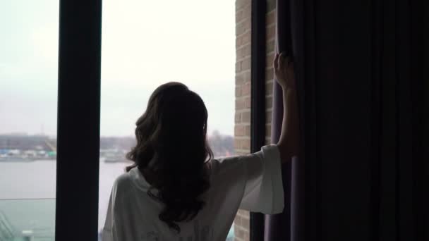 Mujer joven en albornoz de seda sosteniendo zapatos cerca de la silueta de la ventana . — Vídeos de Stock
