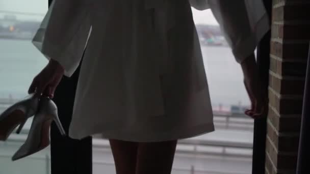 Mujer joven en albornoz de seda sosteniendo zapatos que van a la silueta de la ventana . — Vídeo de stock