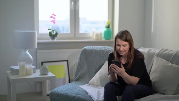 Wanita muda dengan smartphone di sofa di rumah. Perangkat gadget telepon — Stok Video