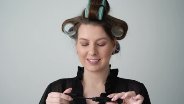 Menina com rolos de encrespadores de cabelo no cabelo, aplicando rímel fazendo maquiagem . — Vídeo de Stock