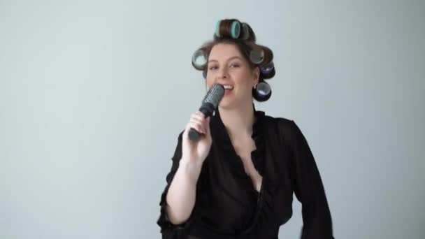 Femme avec des rouleaux de bigoudis sur les cheveux et en peignoir dansant en chantant dans une brosse à cheveux . — Video