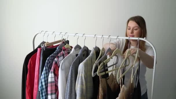 Молодая женщина выбирает одежду на стойке, ищет, что надеть. Магазин или гардероб — стоковое видео