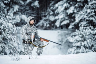 Kamuflaj, erkek avcısı silahlı bir tüfekle bir karlı kış ormanda ayakta