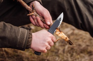 Close-up hand hunter man with knife cut a wooden stick clipart