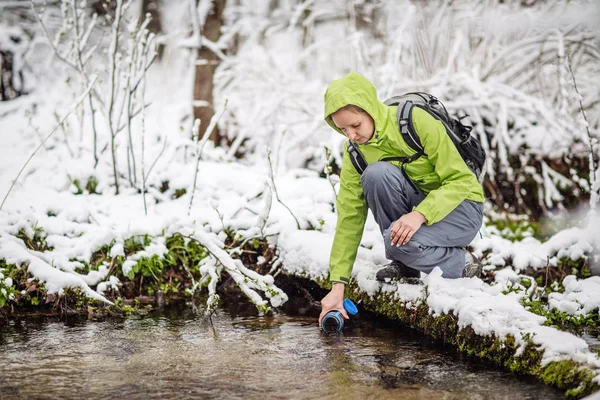 woman filling bottle of water from a winter forest stream. travel and vacation concept