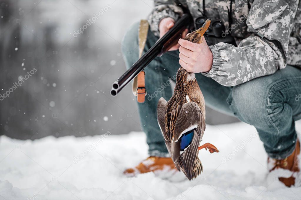 Hunter in camouflage with rifle, holding duck prey  in winter forest. Hunt concept.
