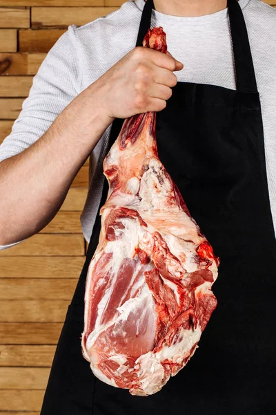 Kasap, eller çiğ et kuzu bacak etini ahşap bac üzerinde tutar — Stok fotoğraf