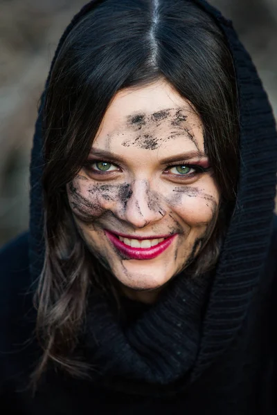 A close up of a woman\'s face and hands covered in mud. Young woman with dirty face and arms on dark background.