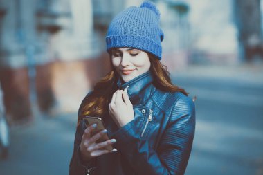 Cep telefonu tutan güzel, şık bir kadın resmi. Genç Avrupalı kız caddede duruyor ve cep telefonu kullanıyor. Kadın telefonla müzik dinliyor ve eğleniyor. Taşınabilir internet konsepti. Sosyal medya.