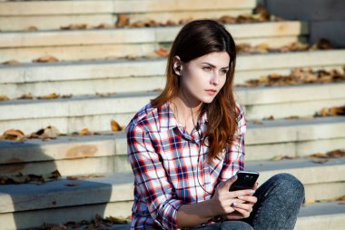 Cep telefonu tutan güzel, şık bir kadın resmi. Genç Avrupalı kız caddede duruyor ve cep telefonu kullanıyor. Kadın telefonla müzik dinliyor ve eğleniyor. Taşınabilir internet konsepti. Sosyal medya.