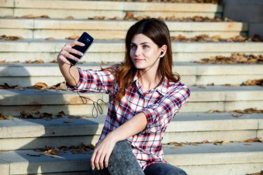 Cep telefonu tutan güzel, şık bir kadın resmi. Genç Avrupalı kız caddede duruyor ve cep telefonu kullanıyor. Kadın telefonla müzik dinliyor ve eğleniyor. Taşınabilir internet konsepti. Sosyal medya.