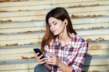 Cep telefonu tutan güzel, şık bir kadın resmi. Genç Avrupalı kız caddede duruyor ve cep telefonu kullanıyor. Kadın telefonla müzik dinliyor ve eğleniyor. Taşınabilir internet konsepti. Sosyal medya.