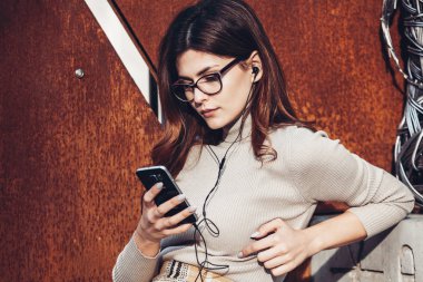 Cep telefonu tutan güzel, şık bir kadın resmi. Genç Avrupalı kız caddede duruyor ve cep telefonu kullanıyor. Kadın telefonla müzik dinliyor ve eğleniyor. Taşınabilir internet konsepti. Sosyal medya.