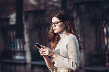 Cep telefonu tutan güzel, şık bir kadın resmi. Genç Avrupalı kız caddede duruyor ve cep telefonu kullanıyor. Kadın telefonla müzik dinliyor ve eğleniyor. Taşınabilir internet konsepti. Sosyal medya.