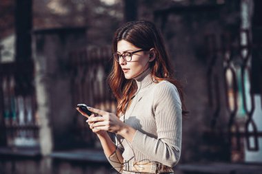 Cep telefonu tutan güzel, şık bir kadın resmi. Genç Avrupalı kız caddede duruyor ve cep telefonu kullanıyor. Kadın telefonla müzik dinliyor ve eğleniyor. Taşınabilir internet konsepti. Sosyal medya.