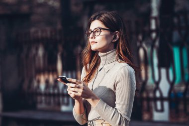 Cep telefonu tutan güzel, şık bir kadın resmi. Genç Avrupalı kız caddede duruyor ve cep telefonu kullanıyor. Kadın telefonla müzik dinliyor ve eğleniyor. Taşınabilir internet konsepti. Sosyal medya.