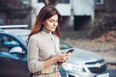 Cep telefonu tutan güzel, şık bir kadın resmi. Genç Avrupalı kız caddede duruyor ve cep telefonu kullanıyor. Kadın telefonla müzik dinliyor ve eğleniyor. Taşınabilir internet konsepti. Sosyal medya.