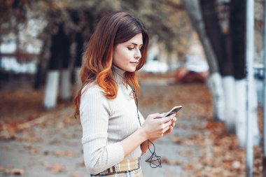 Cep telefonu tutan güzel, şık bir kadın resmi. Genç Avrupalı kız caddede duruyor ve cep telefonu kullanıyor. Kadın telefonla müzik dinliyor ve eğleniyor. Taşınabilir internet konsepti. Sosyal medya.