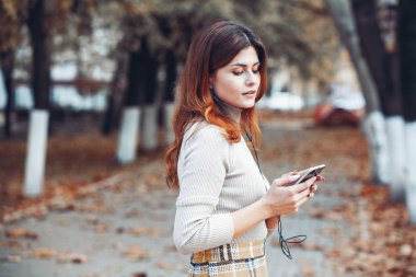 Cep telefonu tutan güzel, şık bir kadın resmi. Genç Avrupalı kız caddede duruyor ve cep telefonu kullanıyor. Kadın telefonla müzik dinliyor ve eğleniyor. Taşınabilir internet konsepti. Sosyal medya.